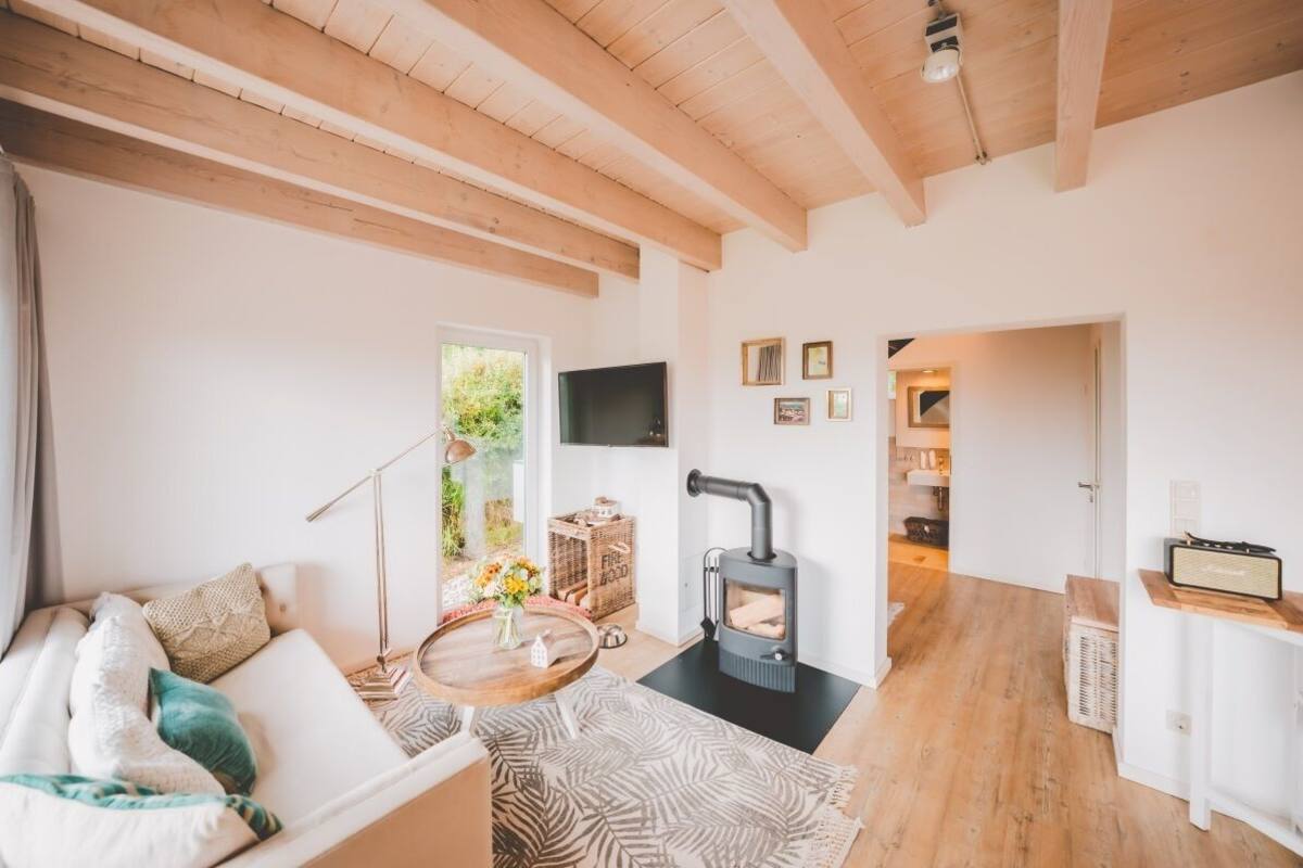A cozy living room with a sofa, a round wooden table, a black wood-burning stove, and a television on the wall.