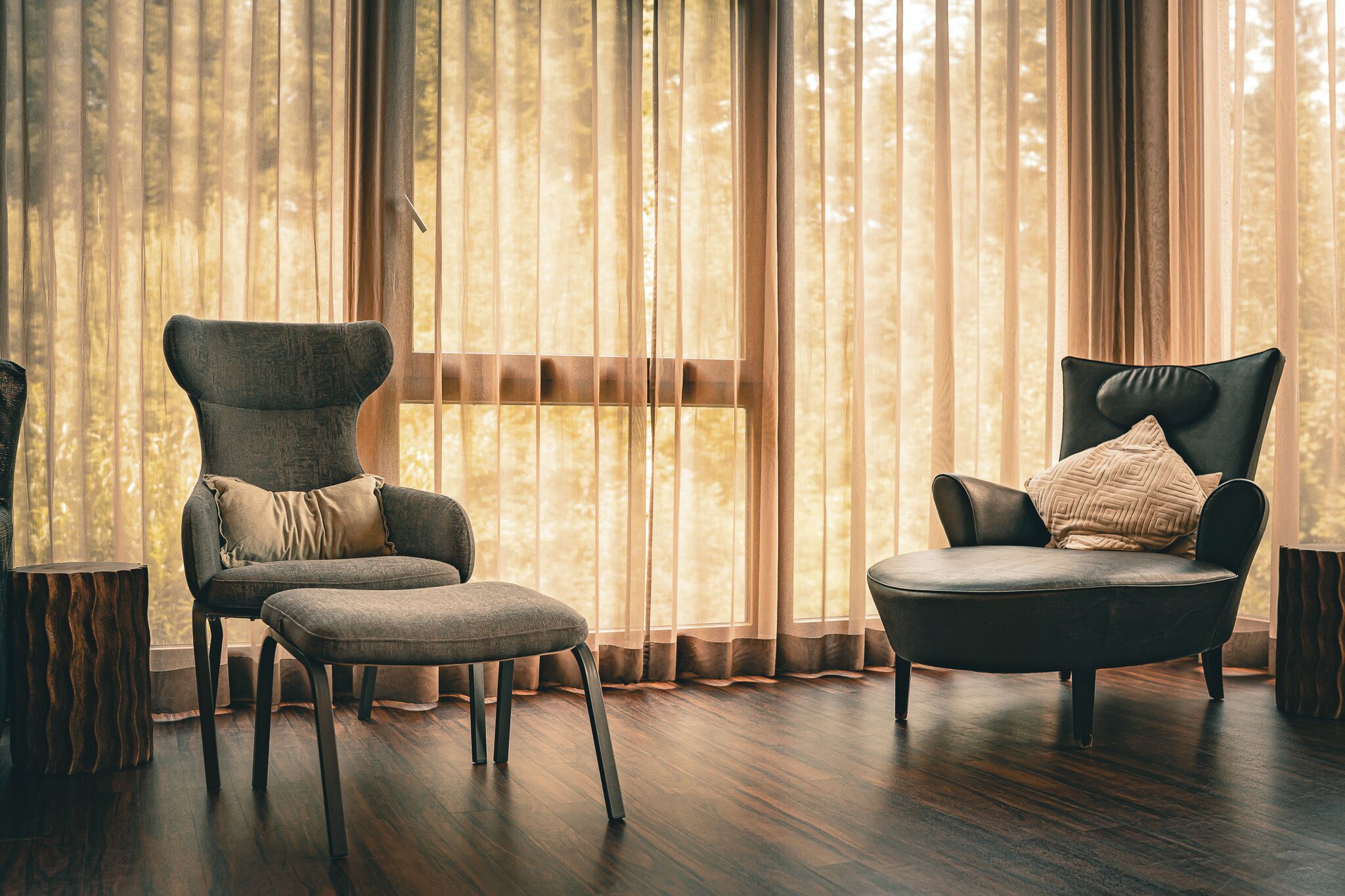A room with two armchairs and a small table, large windows with sheer curtains, and a wooden floor.