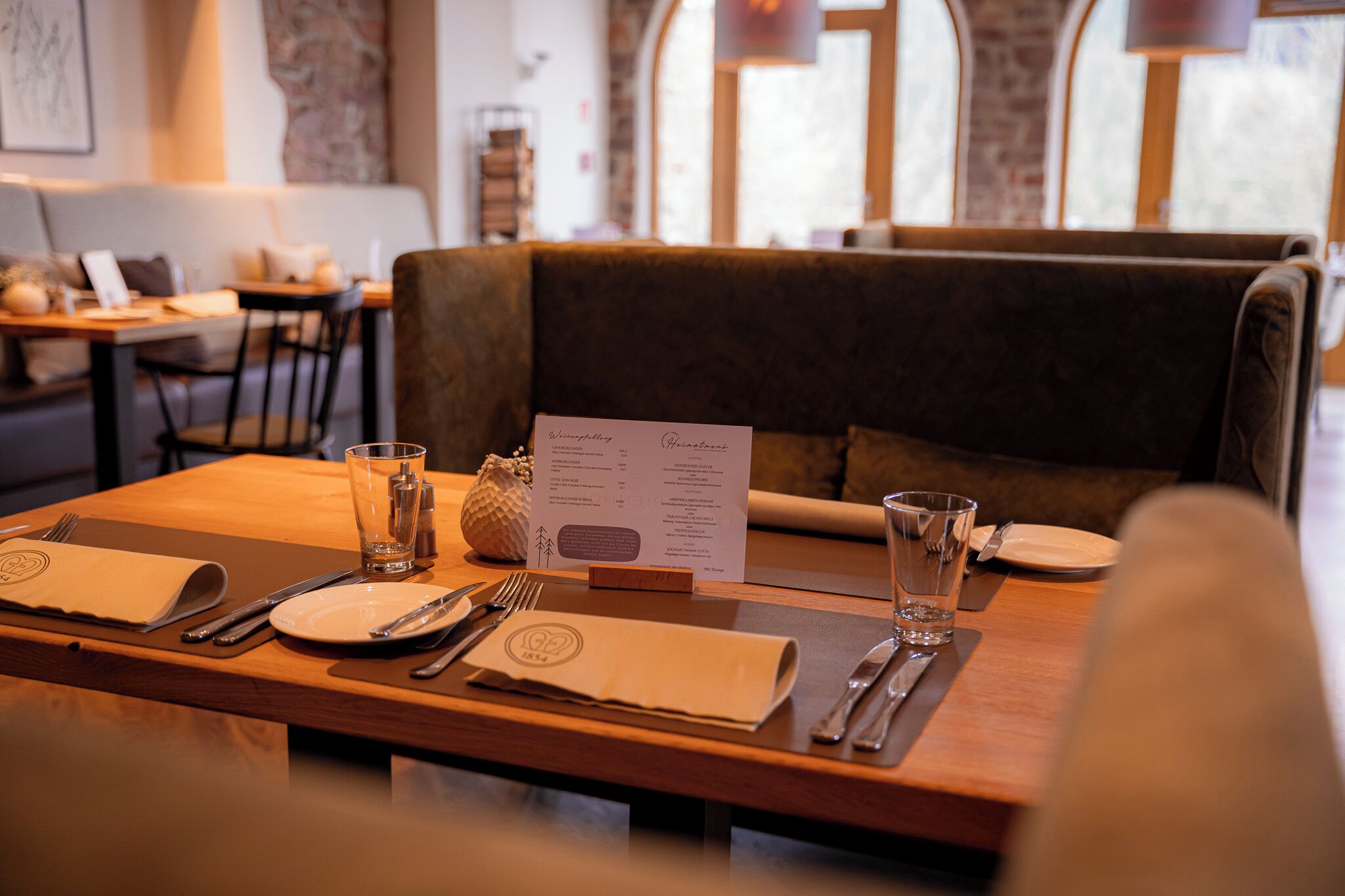 Ein Restaurantambiente mit einem Holztisch, gedeckt für zwei Personen, inklusive Teller, Besteck und Gläsern. Ein Menü liegt auf dem Tisch.