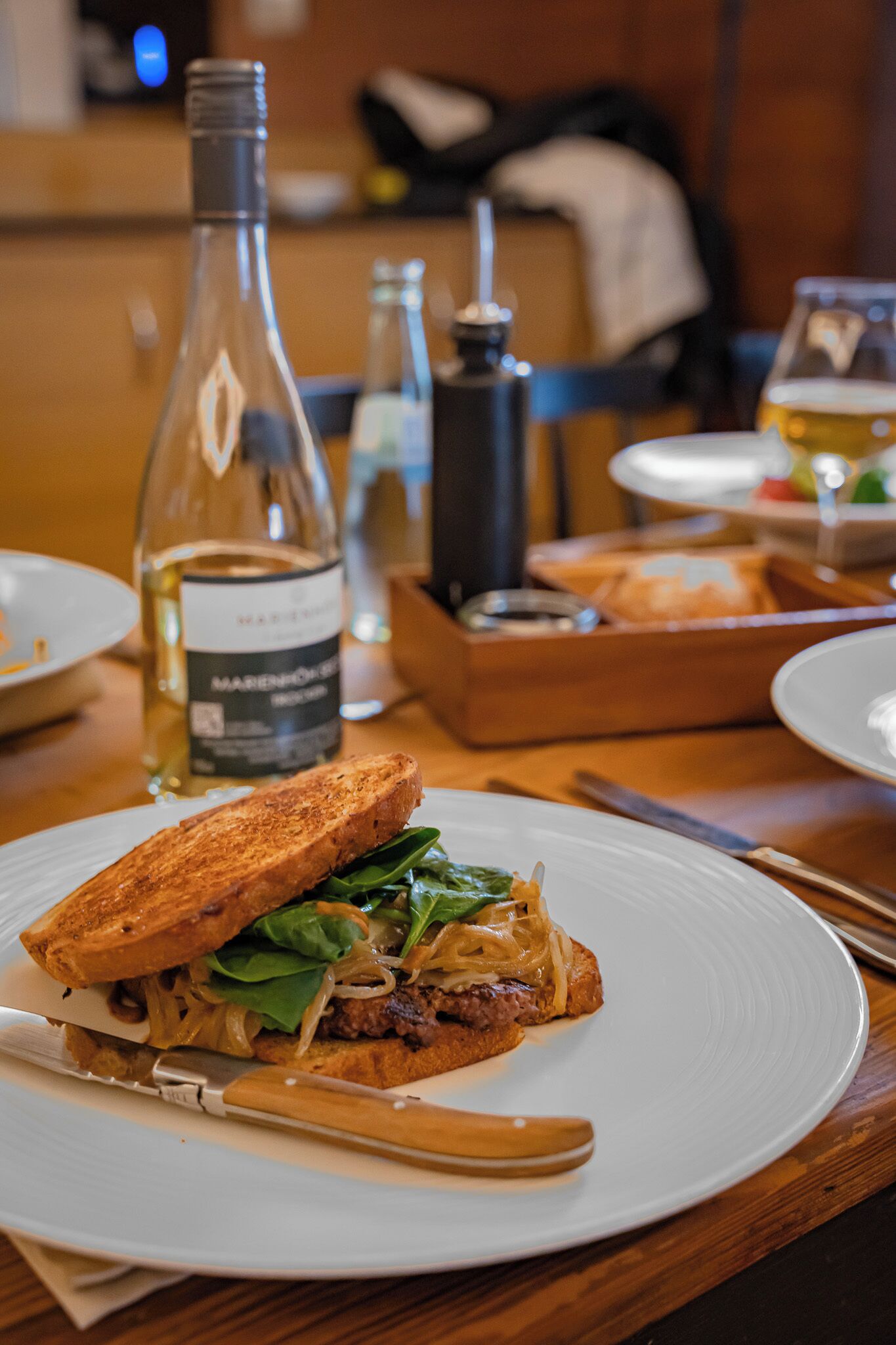 A sandwich on a plate with a knife and a bottle of wine in the background.