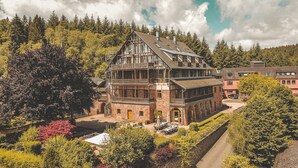 A large, multi-story building with a prominent balcony and a stone facade, surrounded by lush greenery and trees.
