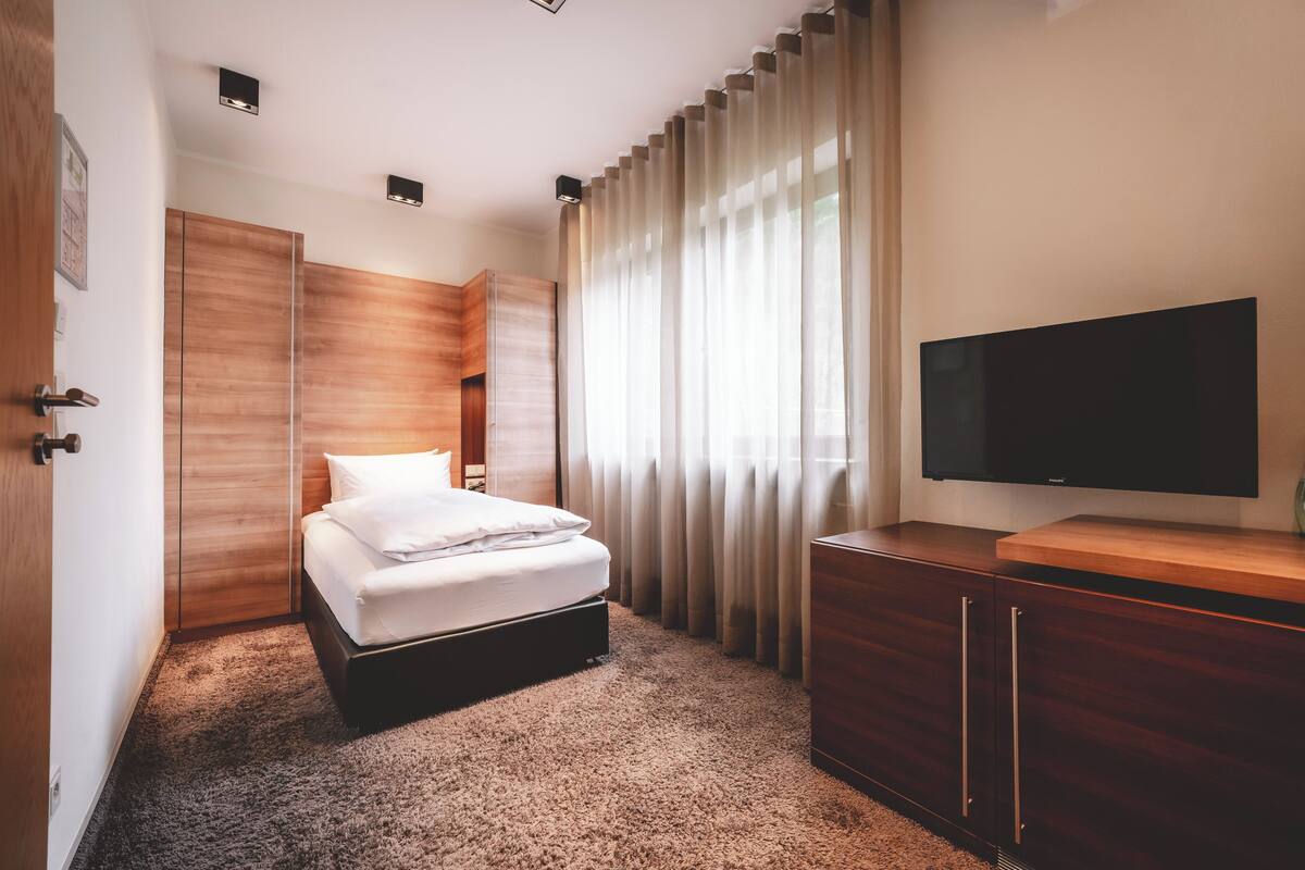 A hotel room with a bed, a TV, a wooden wardrobe, and a large window with curtains.