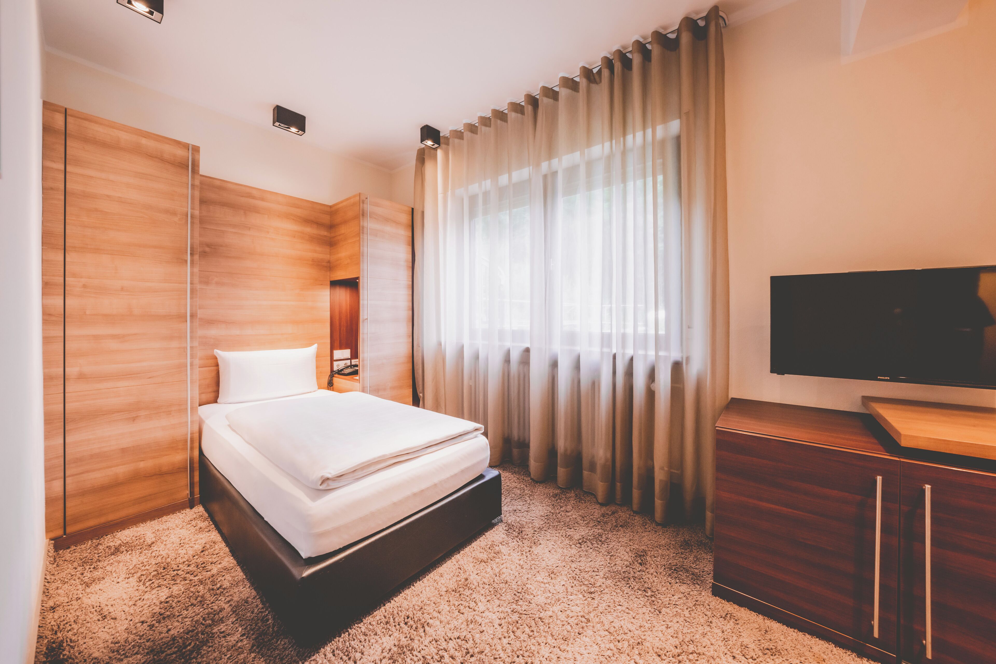 A hotel room with a bed, wooden wardrobe, a TV, and a large window with curtains.