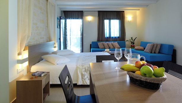 Habitación de hotel con cama, sofá azul, mesa de comedor con fruta y copas de vino, y aire acondicionado de pared.