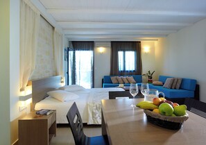 A hotel room with a bed, a desk, a dining table, a blue sofa, and a fruit basket. - Minos Village (Chania)