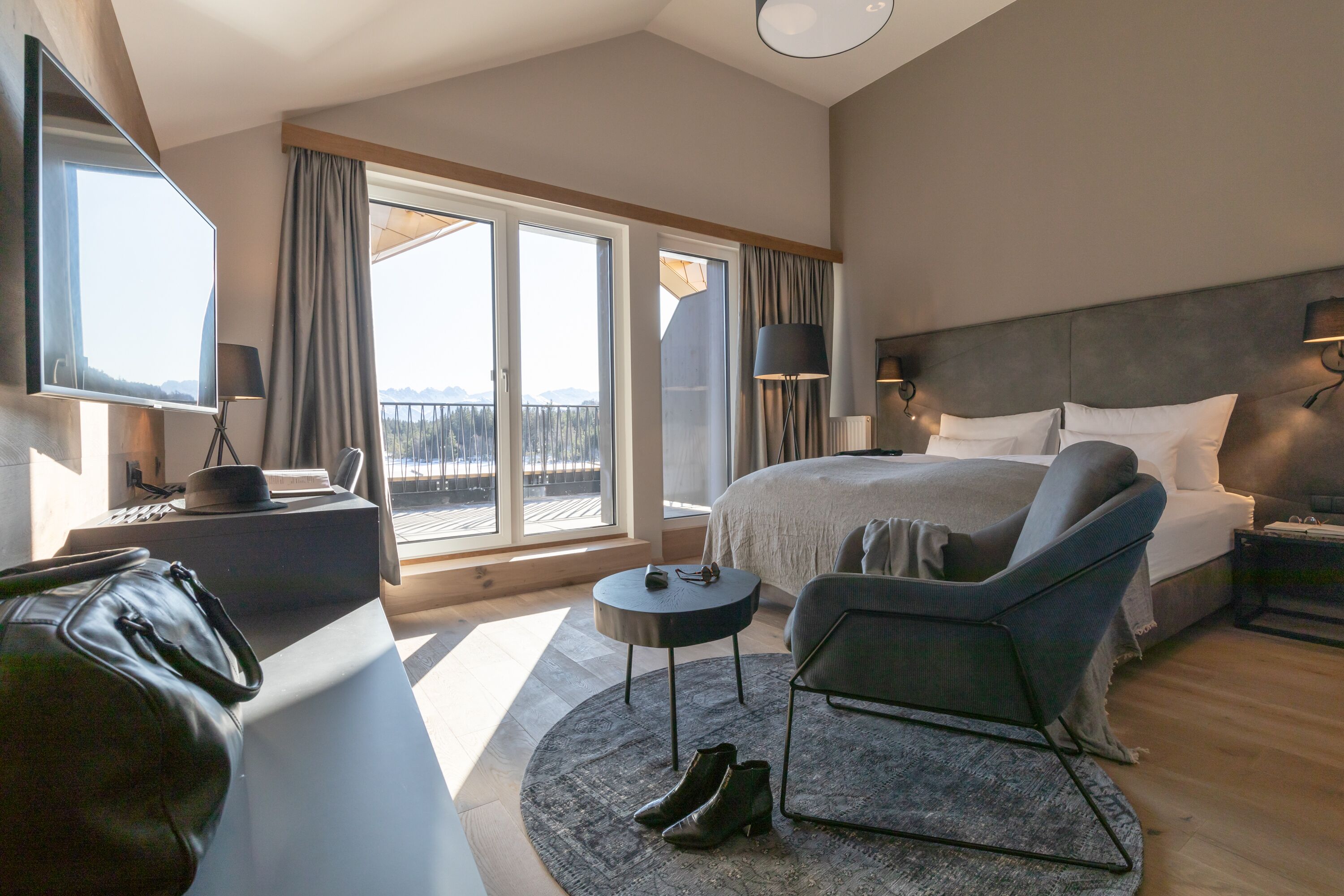 A modern hotel room with a large bed, a seating area with a chair and a small table, a TV, and a view of the outdoors through large windows.