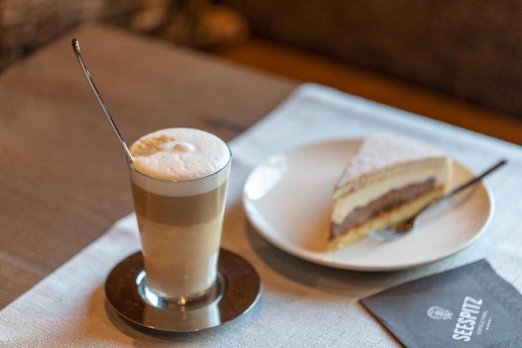 A tall glass of a layered beverage with a spoon, a slice of cake on a plate, and a menu on a table.