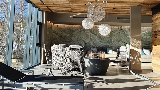 Een modern interieur met een houten plafond, grote ramen en een zithoek met rieten stoelen en een zwart poef.
