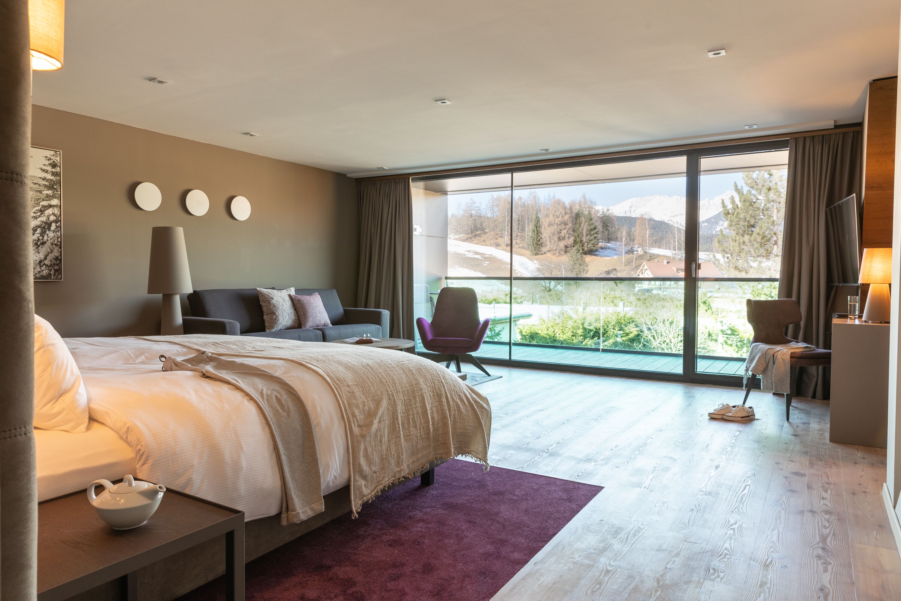 A modern bedroom with a large bed, a seating area with a chair, a small table, and a view of a mountainous landscape through large windows.