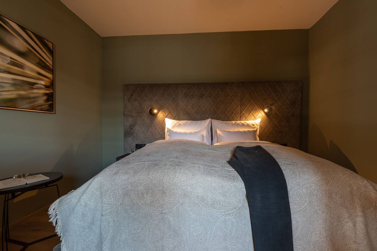 Ein ordentlich bezogenes Bett mit Kopfteil, ein Nachttisch mit Lampe und ein Gemälde an der Wand.