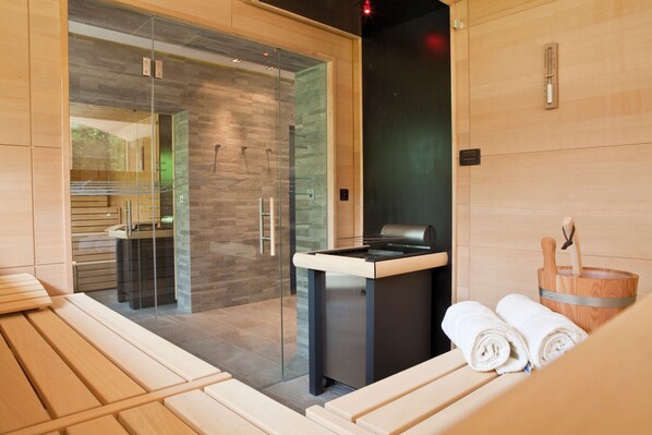 A modern sauna with glass walls, wooden benches, and a wooden bucket with towels. - Hotel Seespitz Seefeld Superior - Adults & Residents Only (Seefeld in Tirol)
