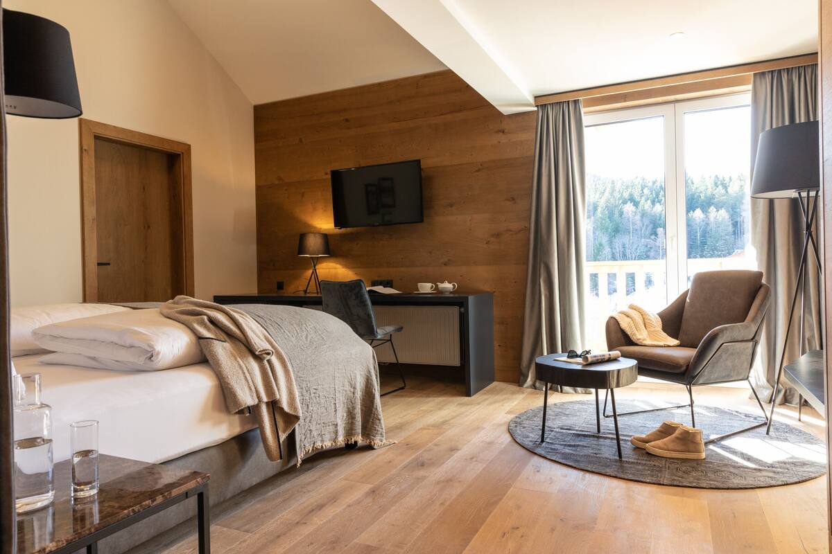 Ein modernes Hotelzimmer mit Bett, Schreibtisch, Stuhl und einem großen Fenster mit Blick auf Bäume.