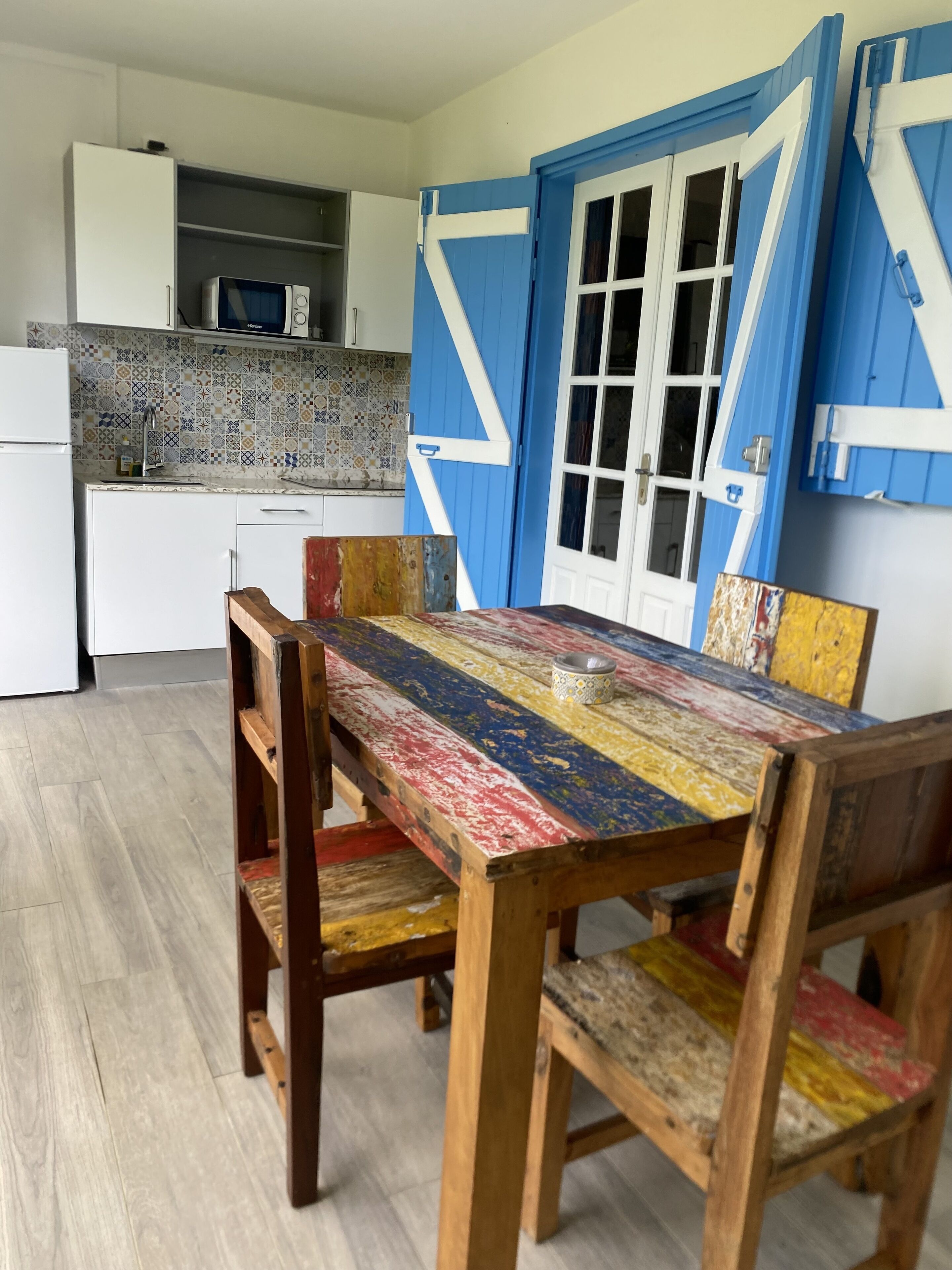 A dining area with a wooden table and chairs, a kitchen in the background, and a blue door with white trim.
