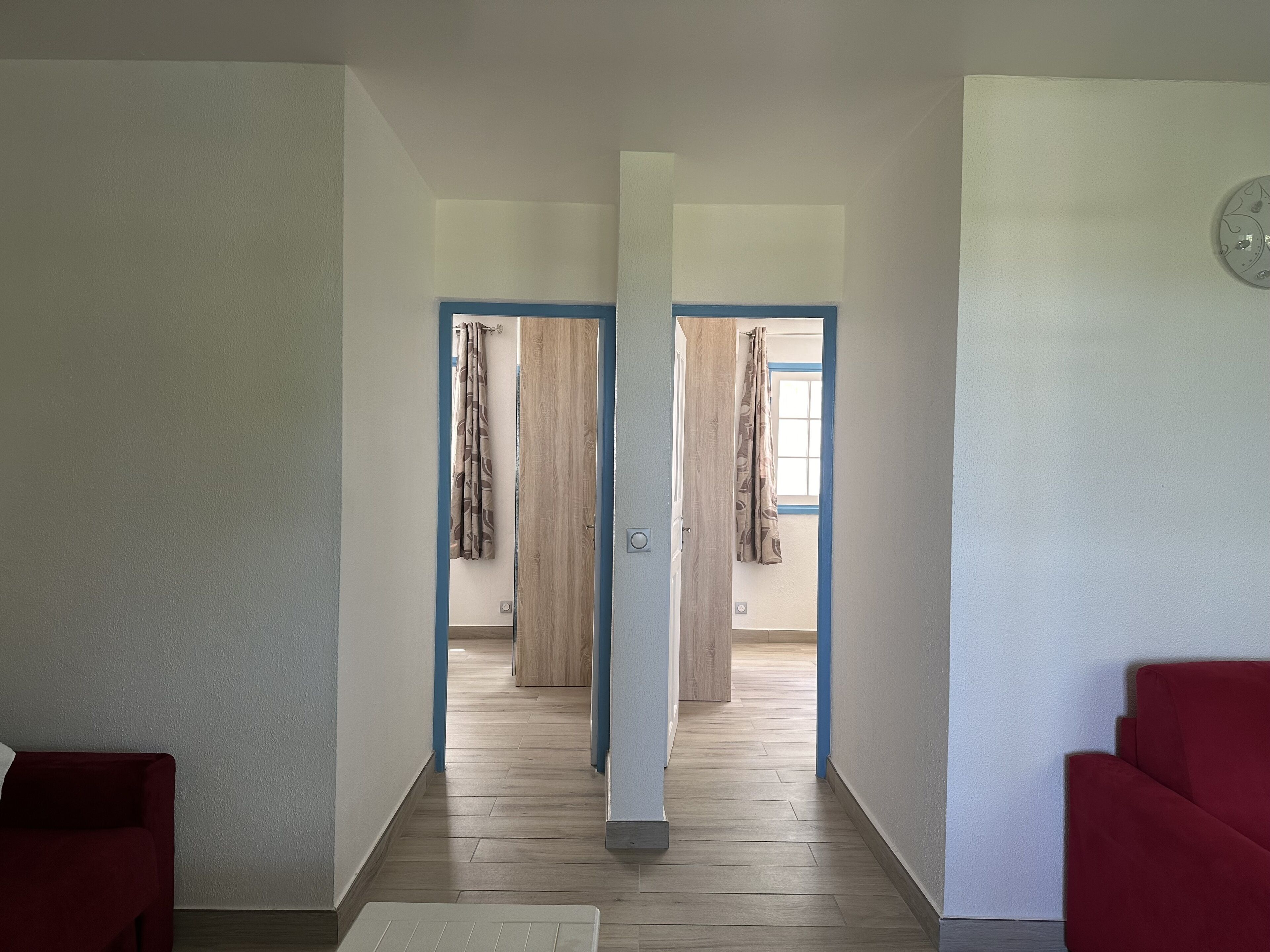 A hallway with a wooden floor, a red sofa on the left, and a red sofa on the right. There are two doors with curtains in the background.