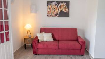 A red sofa with patterned pillows, a wall-mounted zebra painting, a small wooden side table with a lamp, and a white door with red curtains.