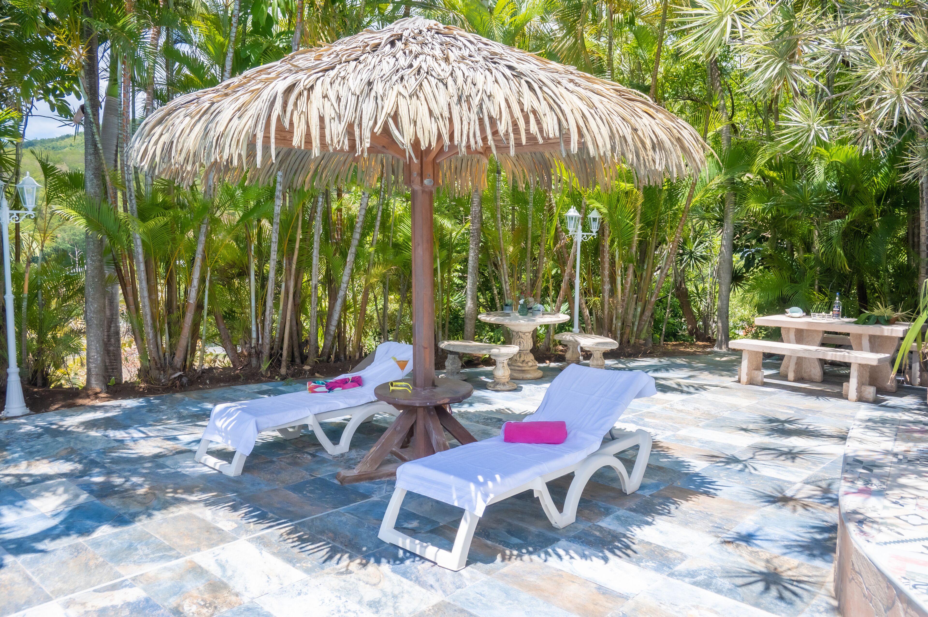 A sunlit outdoor lounge area with two white lounge chairs, a thatched umbrella, and a stone fountain.