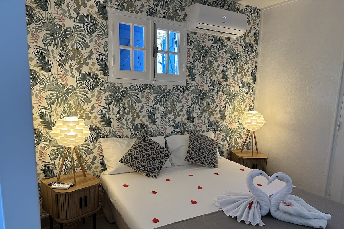 Une chambre à coucher comprenant un lit, deux tables de chevet, un climatiseur fixé au mur et une fenêtre avec des volets bleus.