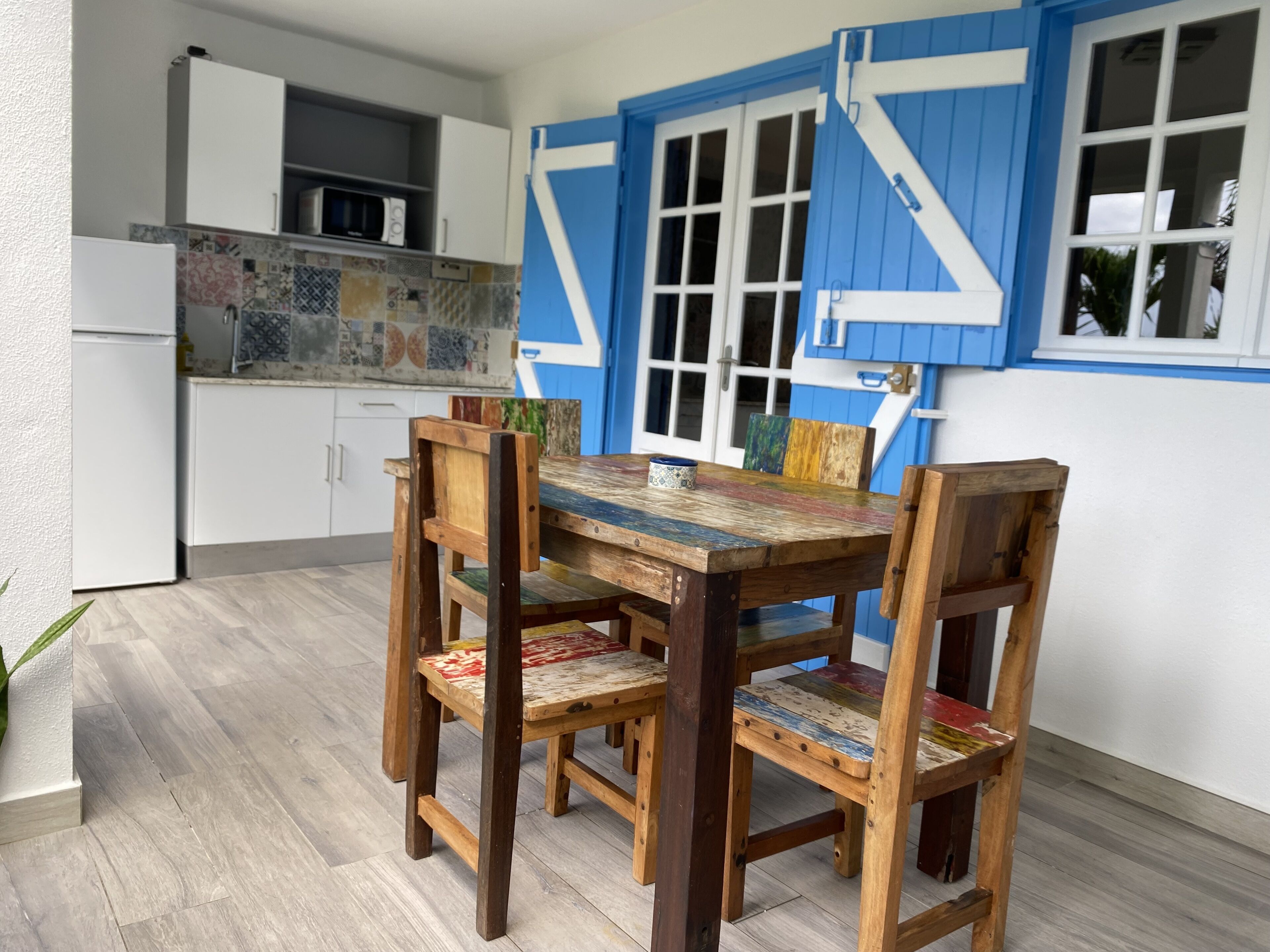Un comedor con una mesa y sillas de madera, una cocina al fondo y una pared exterior azul y blanca.