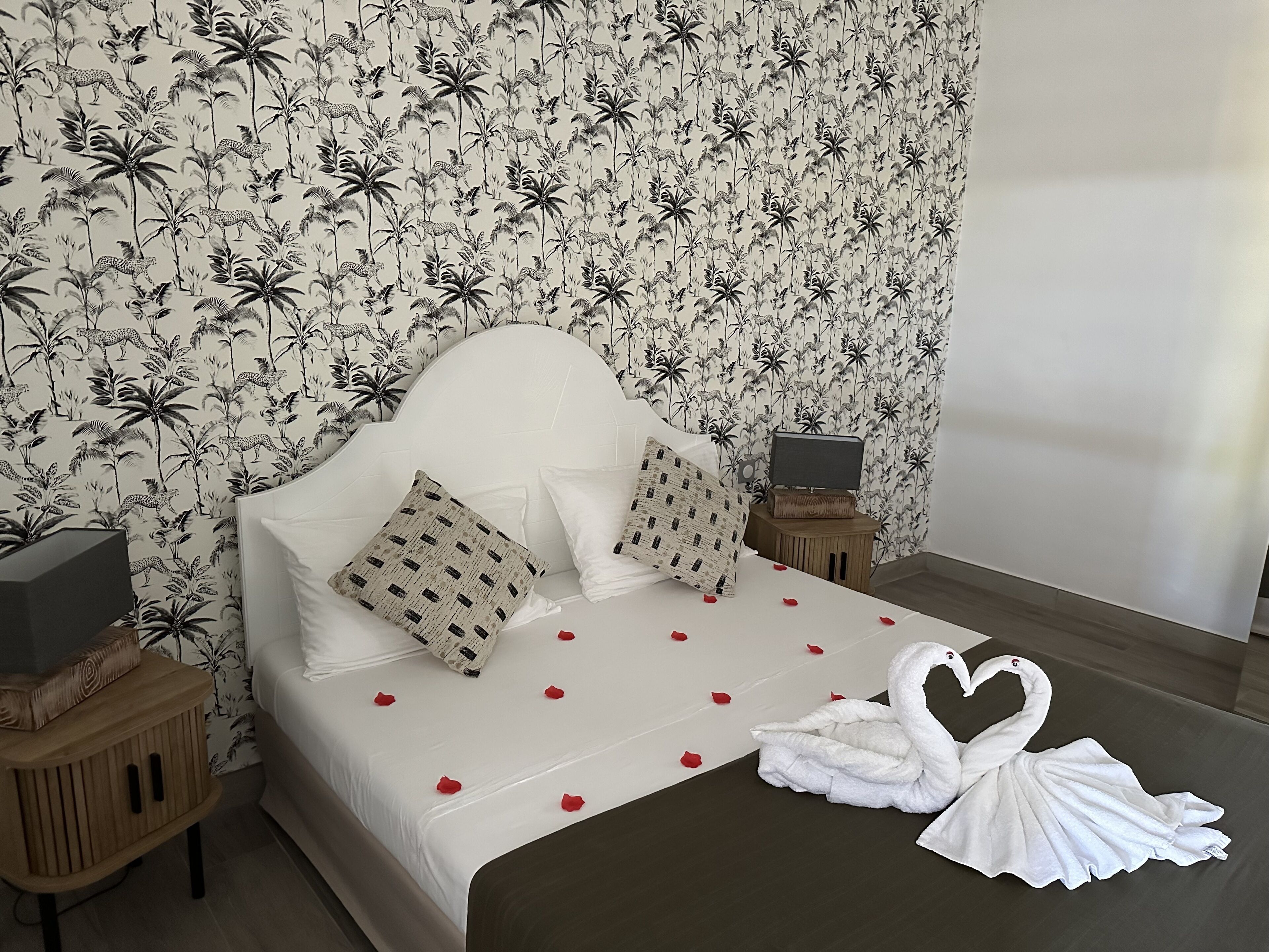 A neatly made bed with a white headboard, two pillows, and a swan-shaped towel arrangement on a bedspread with rose petals.