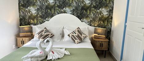 A neatly made bed with a green bedspread and two pillows, a wall with a tropical leaf pattern, and two bedside lamps on wooden nightstands.