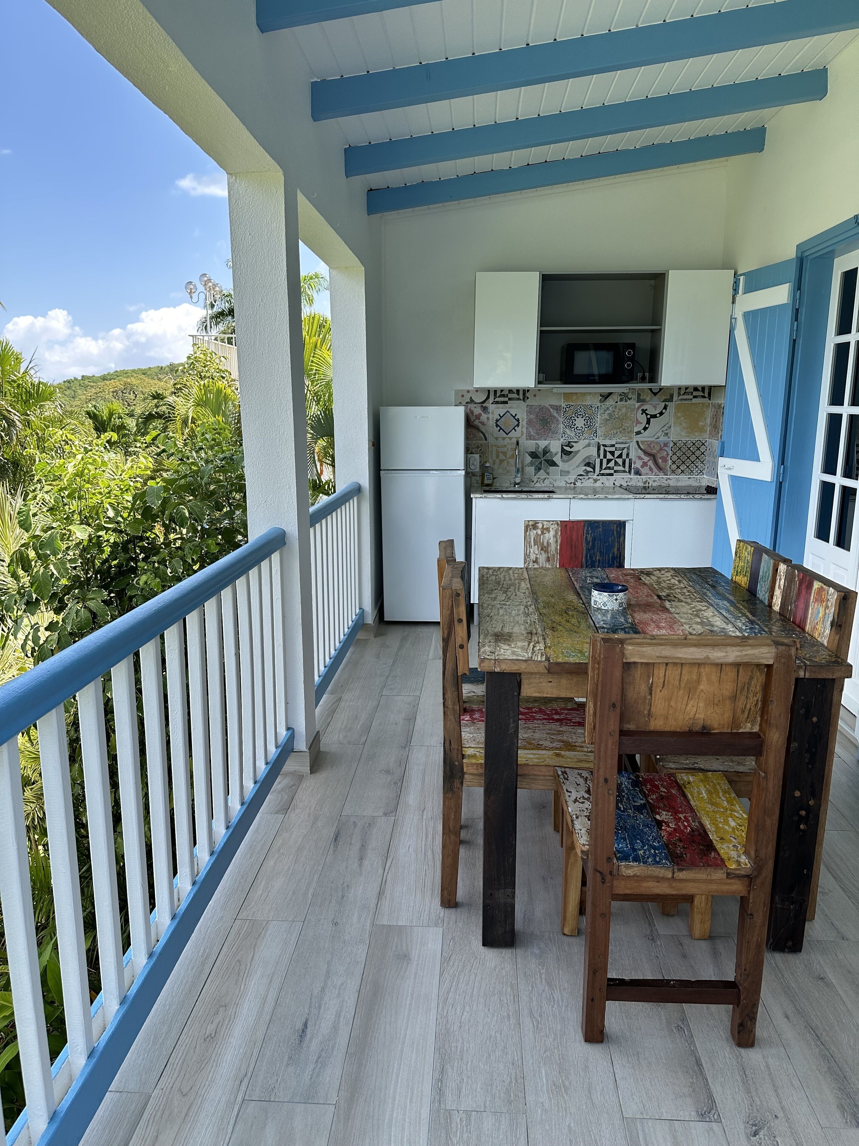 A covered outdoor dining area with a wooden table and chairs, a white refrigerator, and a microwave above a tiled backsplash.