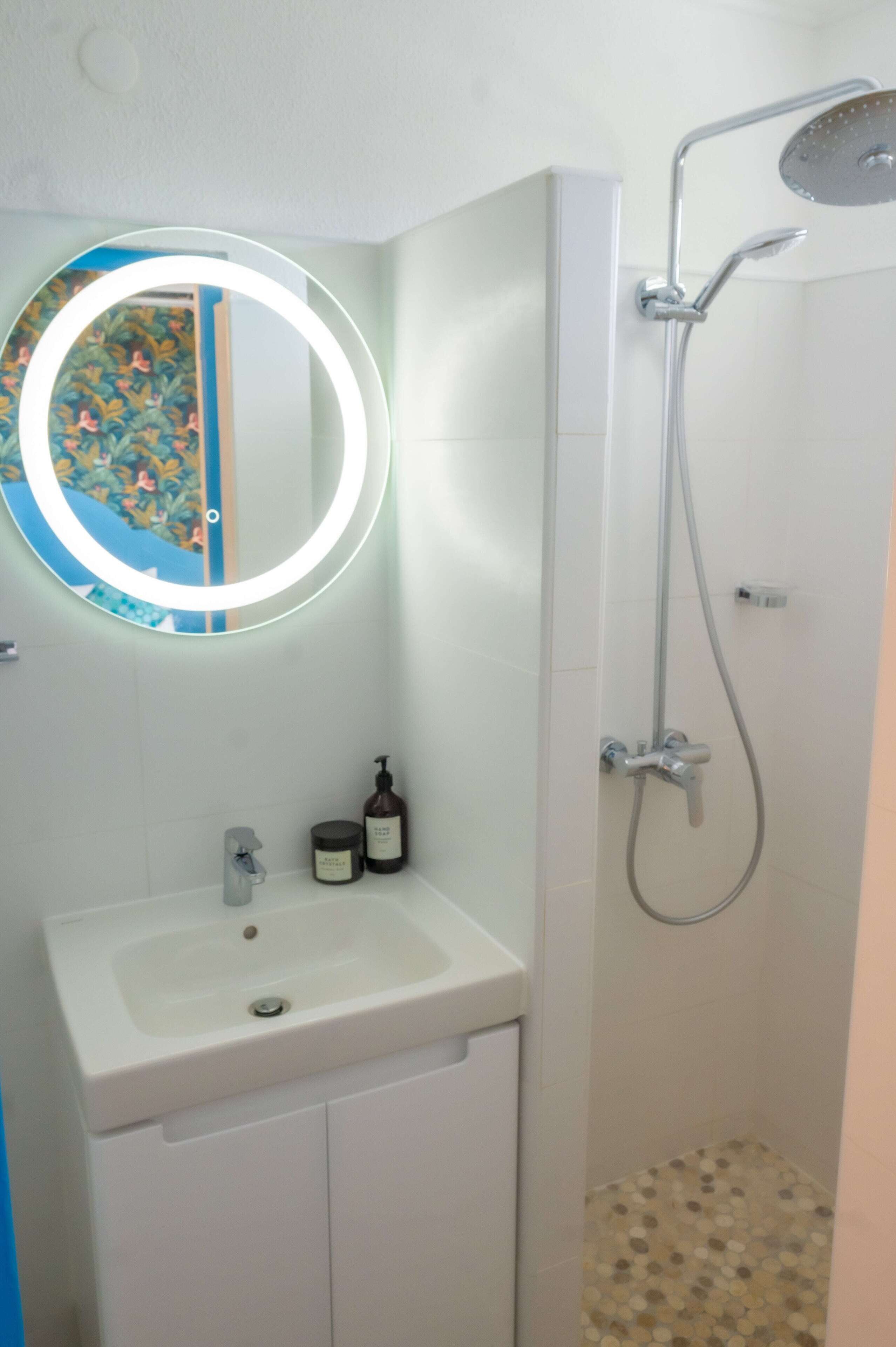 Une salle de bain avec un lavabo blanc, un miroir rond avec un éclairage intégré et une douche.