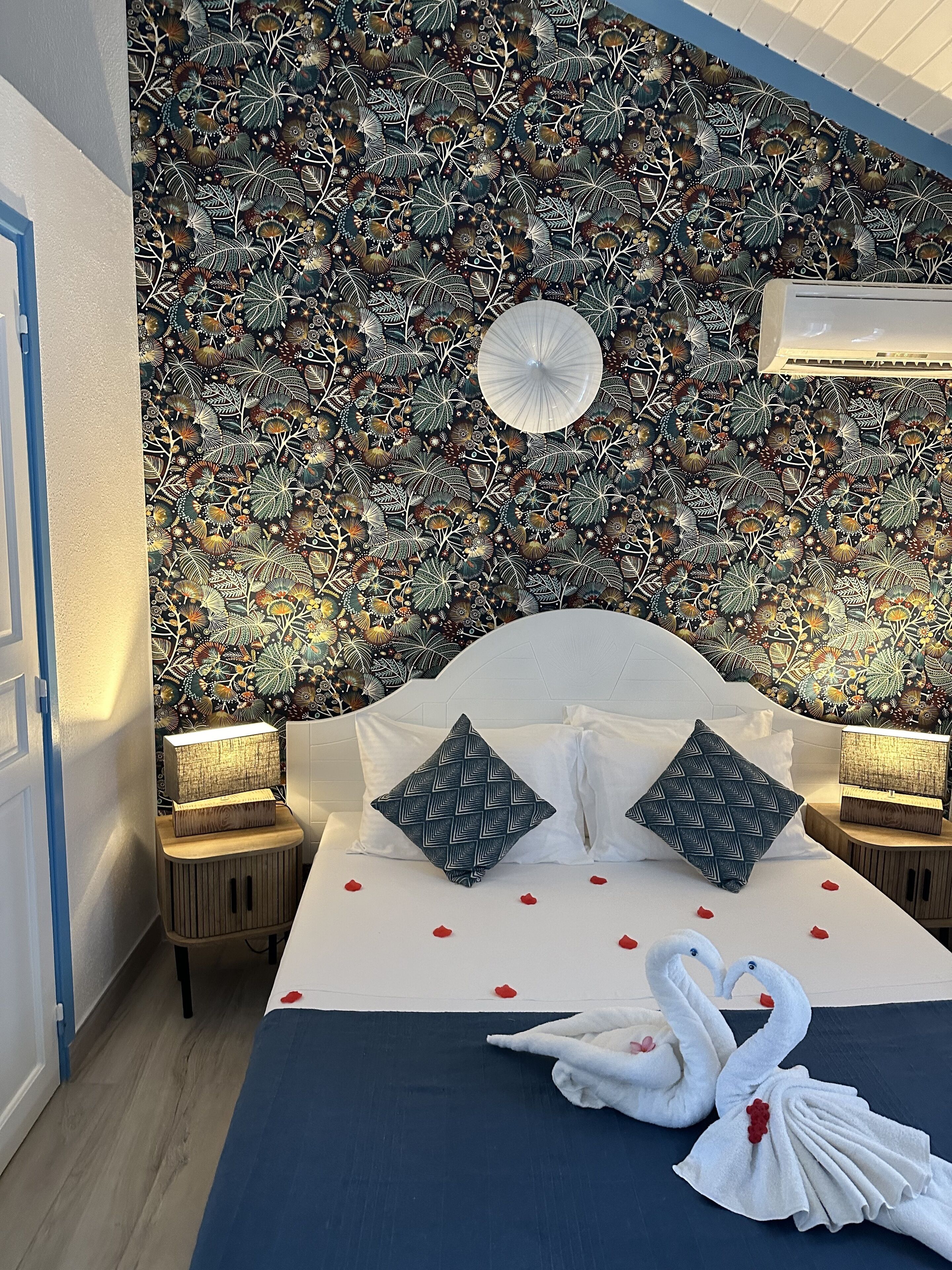 Een slaapkamer met een patroonbehang, een bed met witte beddengoed en decoratieve kussens, en een zwaan-handdoek creatie.