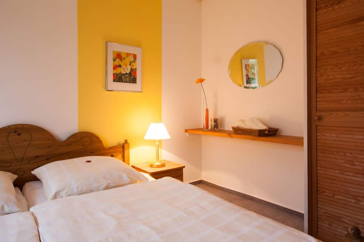 A hotel room with a bed, a bedside table with a lamp, a mirror, a shelf with a vase and a picture, and a wooden headboard.