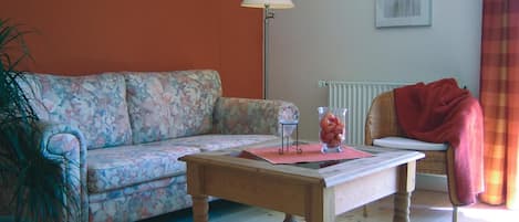 A living room with a floral sofa, a wooden coffee table, a floor lamp, a wicker chair, and a radiator.