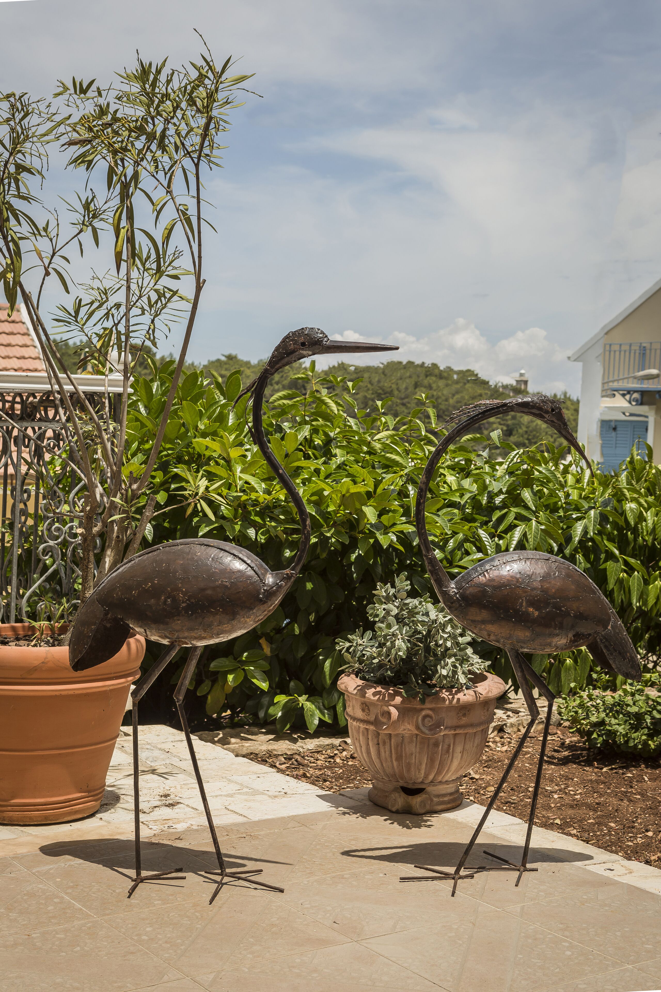 Two metal sculptures of birds in a garden setting with potted plants and a tiled patio.