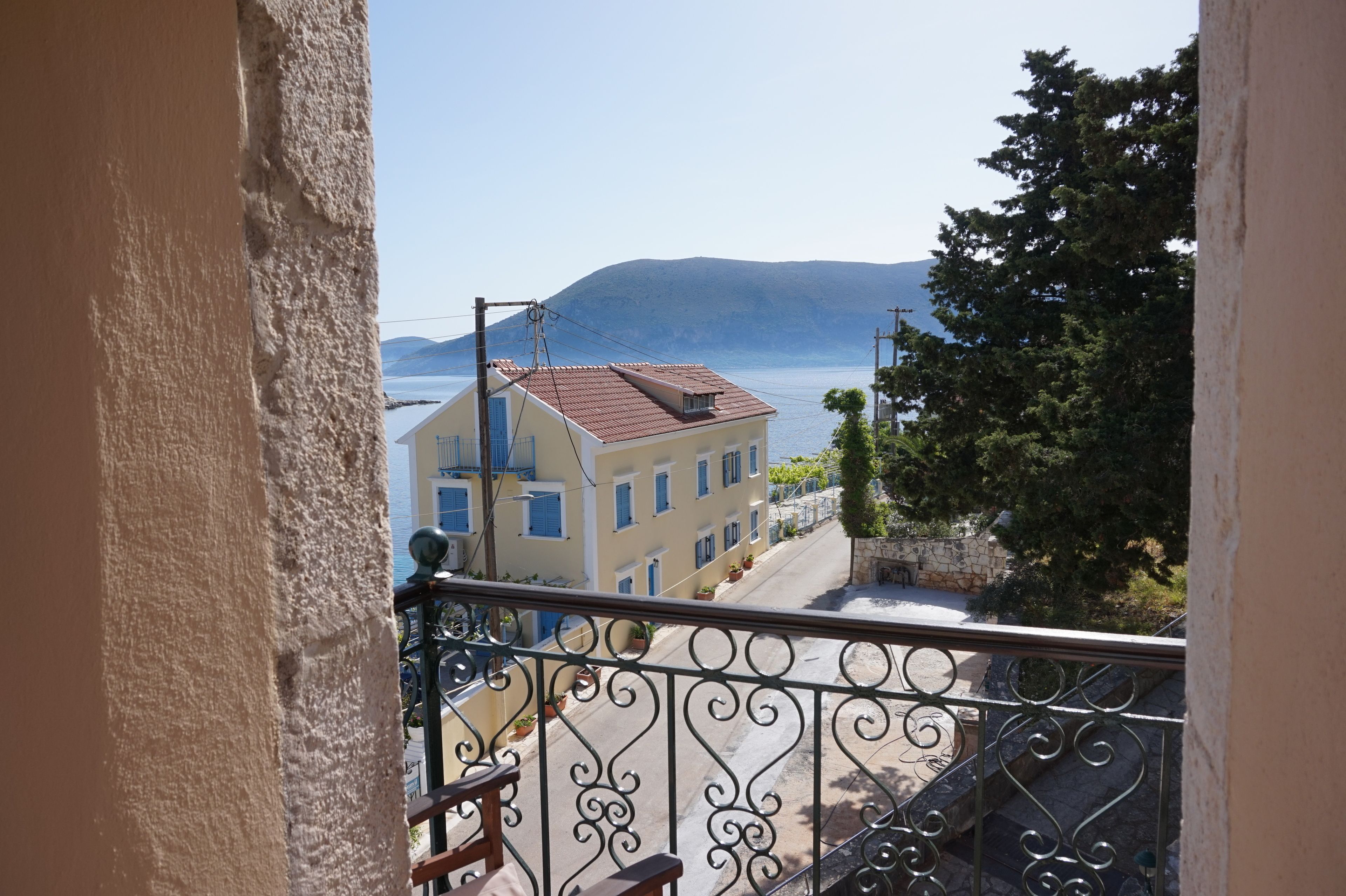 A balcony with a wrought iron railing overlooking a street with buildings and a body of water in the distance.