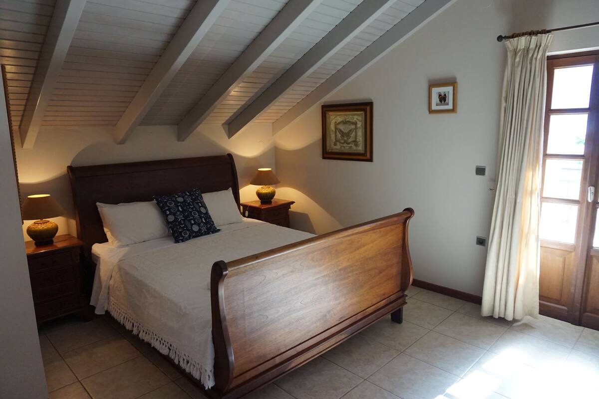 A bedroom with a wooden bed, two bedside tables with lamps, a framed picture on the wall, and a window with curtains.