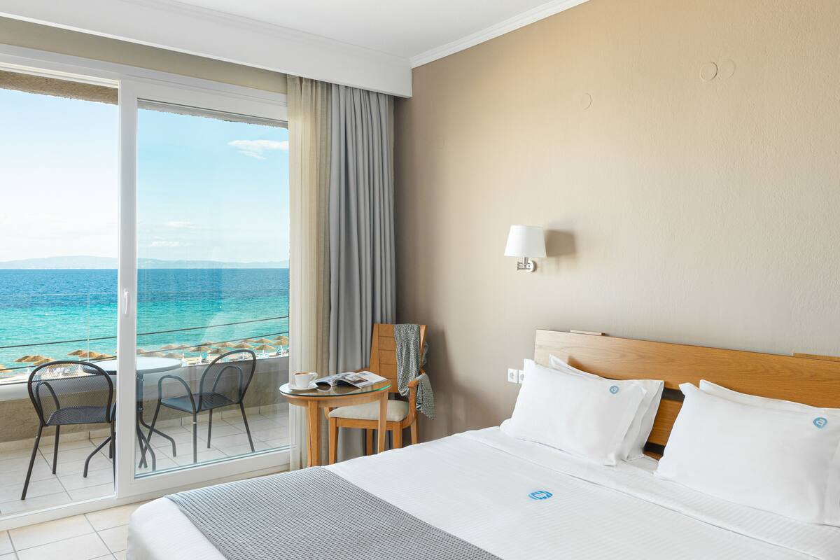Habitación de hotel con cama, una mesita con una silla, una lámpara y vista al océano a través de una puerta corrediza de vidrio.