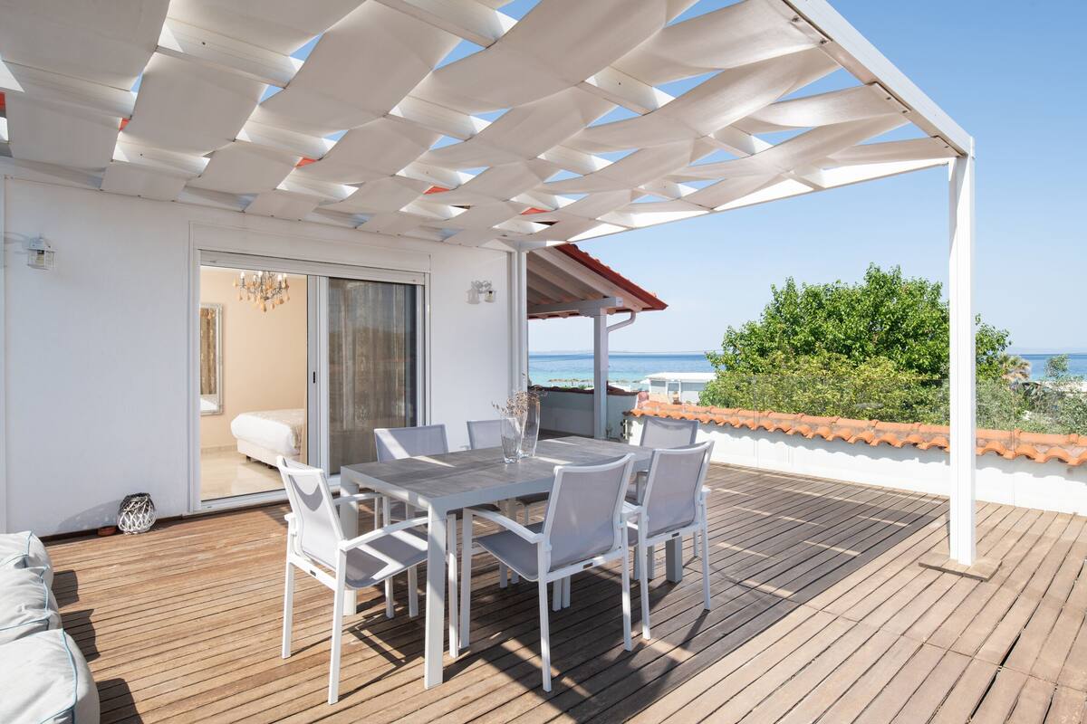 Una terraza de madera con una pérgola blanca, una mesa de comedor con sillas y vista al mar.