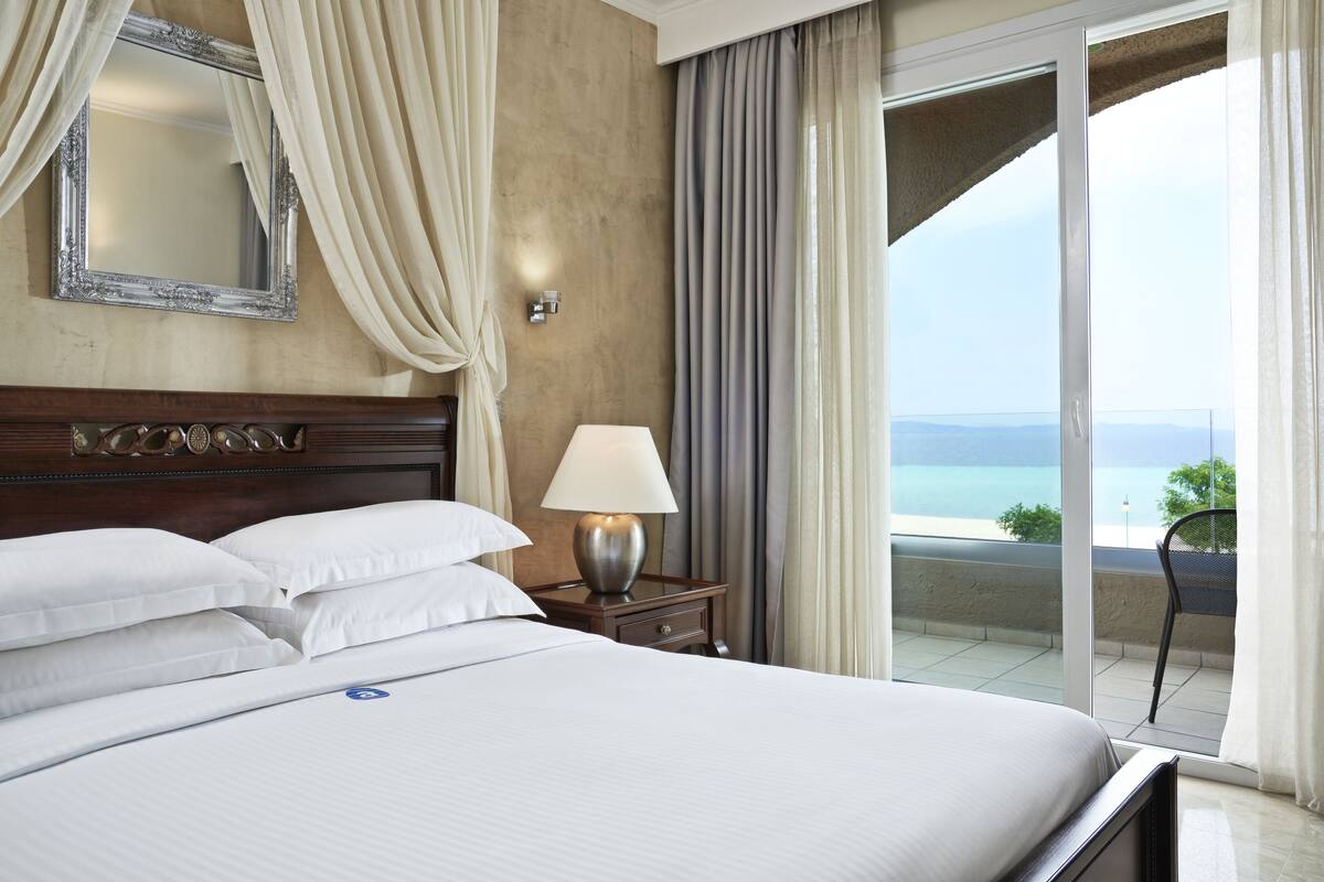 Habitación de hotel con una cama grande, una mesita de noche con lámpara, un espejo y vista al mar a través de una puerta corrediza de vidrio.
