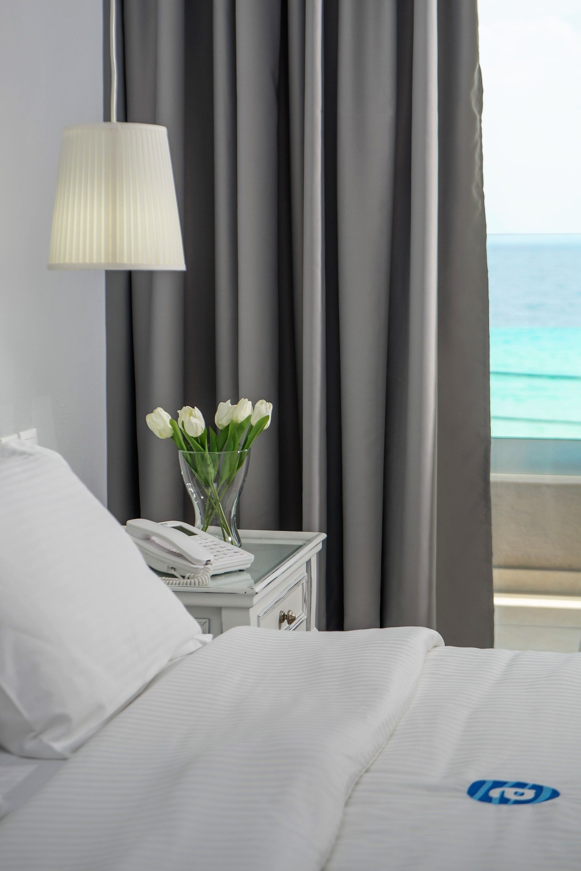 A hotel room with a bed, a bedside table with a phone, a vase with white flowers, and a view of the sea through curtains.