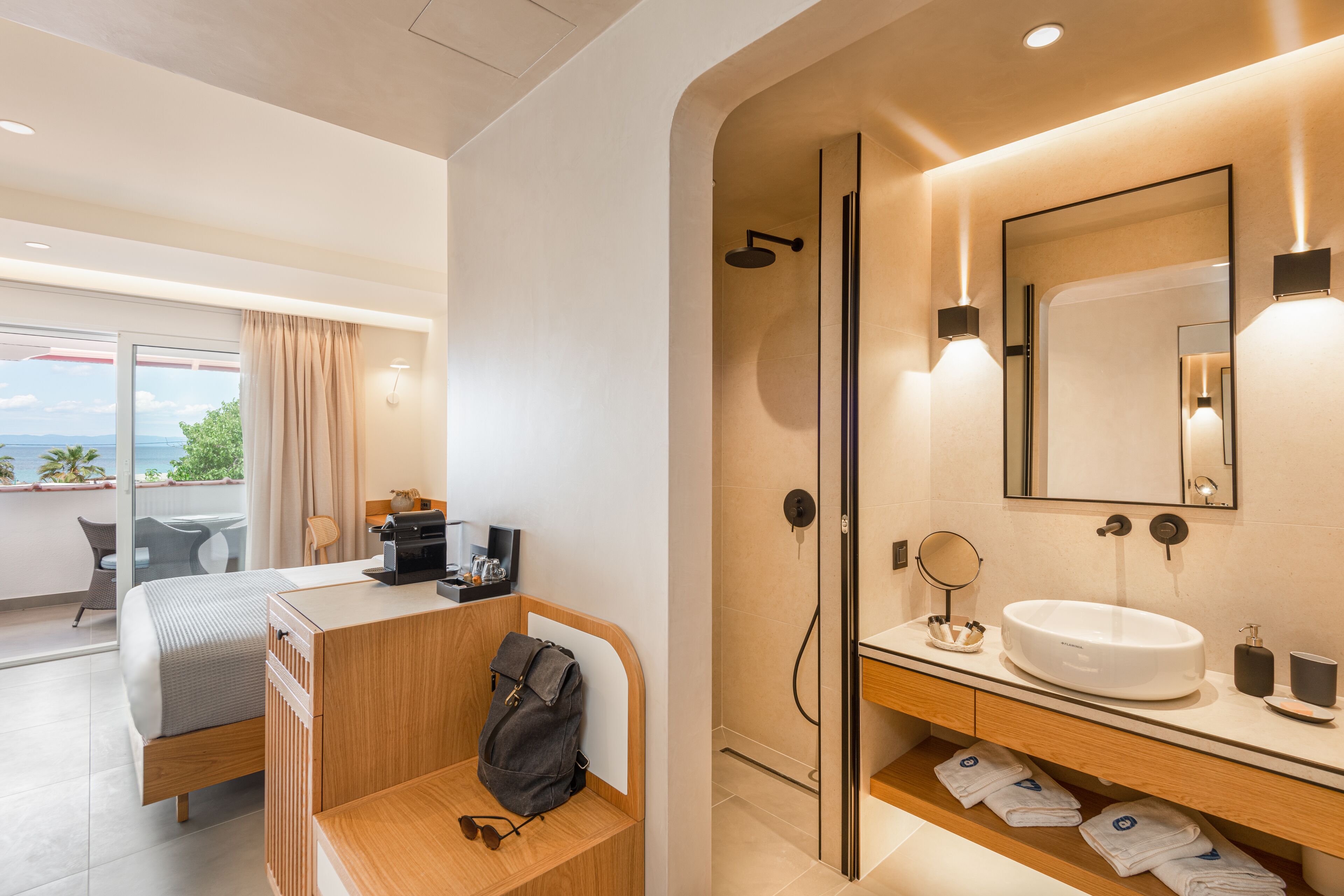 A modern hotel room with a bed, a desk with a laptop, a shower area, and a bathroom with a sink and mirror.