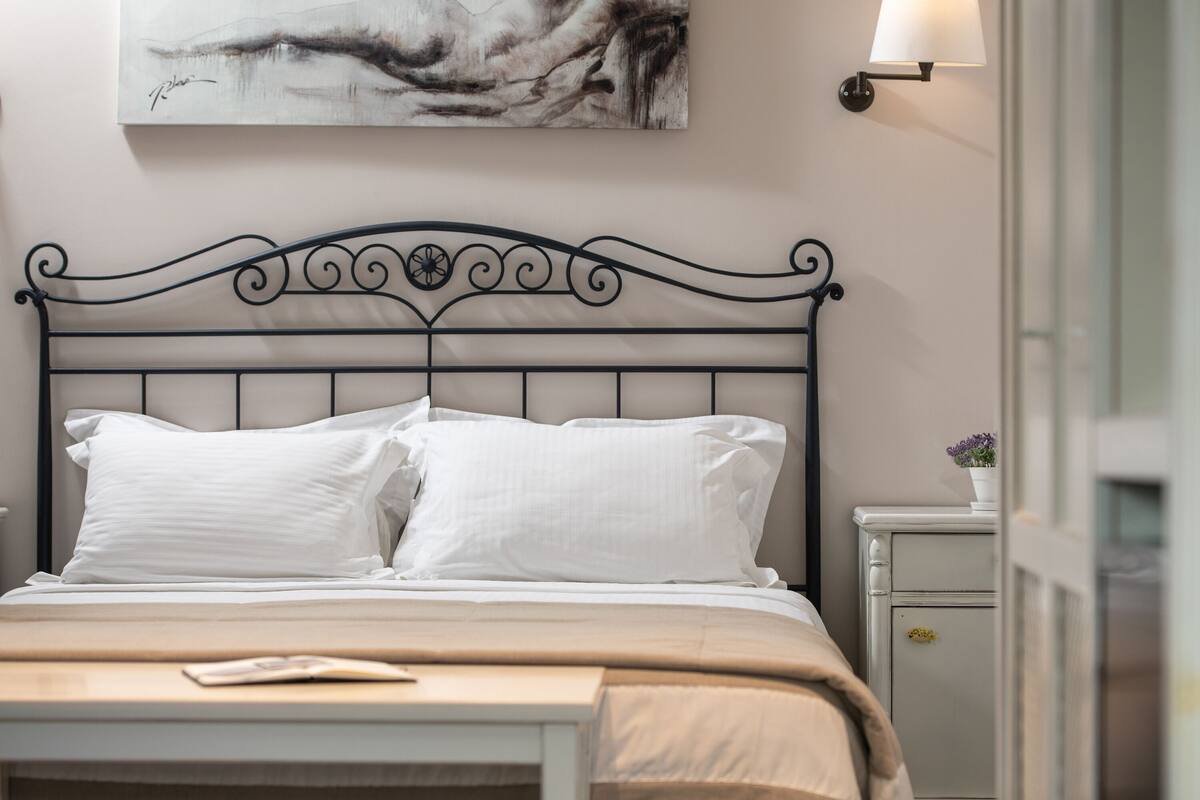 Una cama bien hecha con cabecera de metal, una mesita de noche con lámpara y un cuadro en la pared.