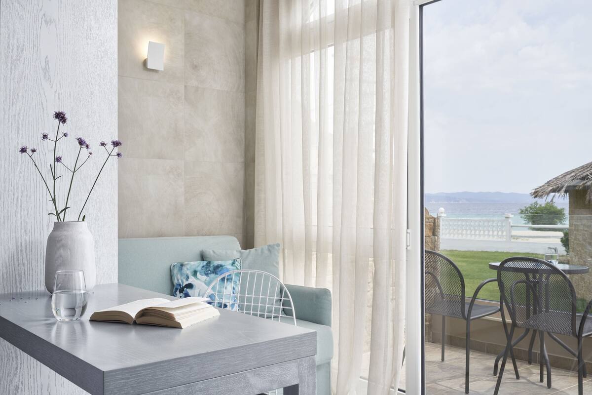Habitación moderna con pared con textura de madera, un florero blanco con flores moradas, un vaso de vidrio transparente y un libro abierto sobre una mesa gris. Un sofá azul claro con cojines estampados se encuentra frente a una ventana grande con cortinas translúcidas, con vistas a un área verde y una valla blanca.