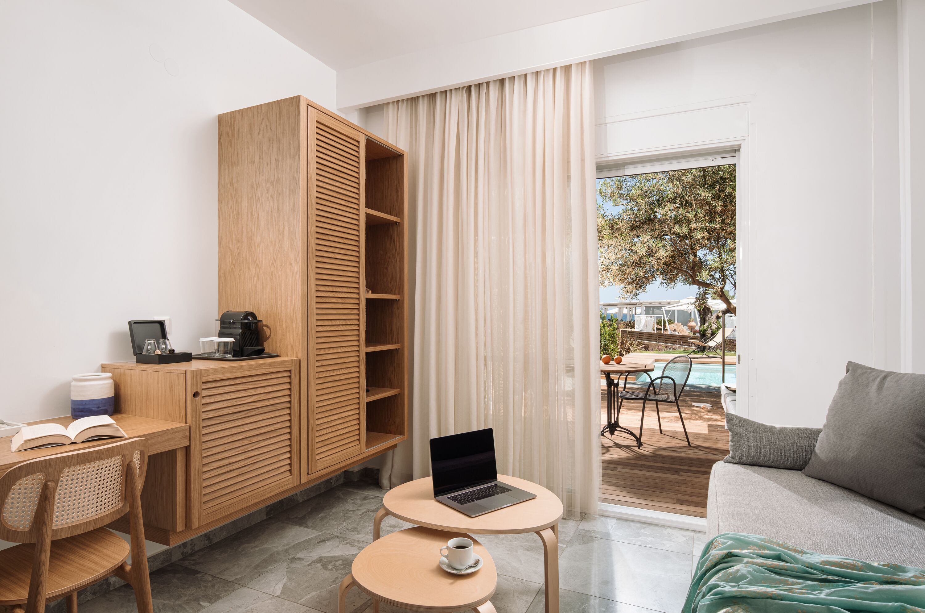 A modern room with a wooden desk, a laptop, a coffee machine, and a view of an outdoor pool area.