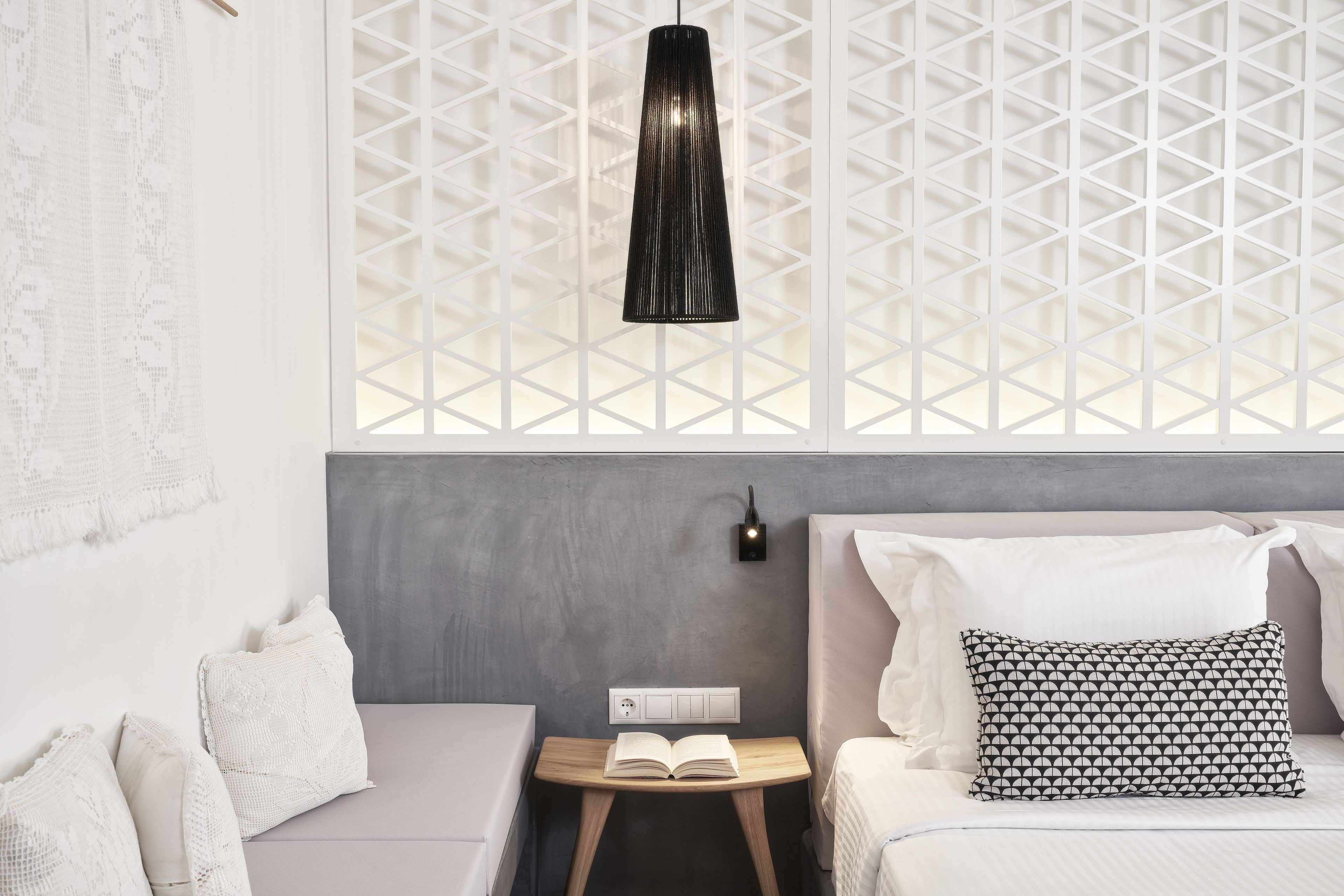 Une chambre moderne avec un mur gris, un lit blanc agrémenté d’oreillers à motifs, une table de chevet sur laquelle repose un livre ouvert et une lampe suspendue noire.