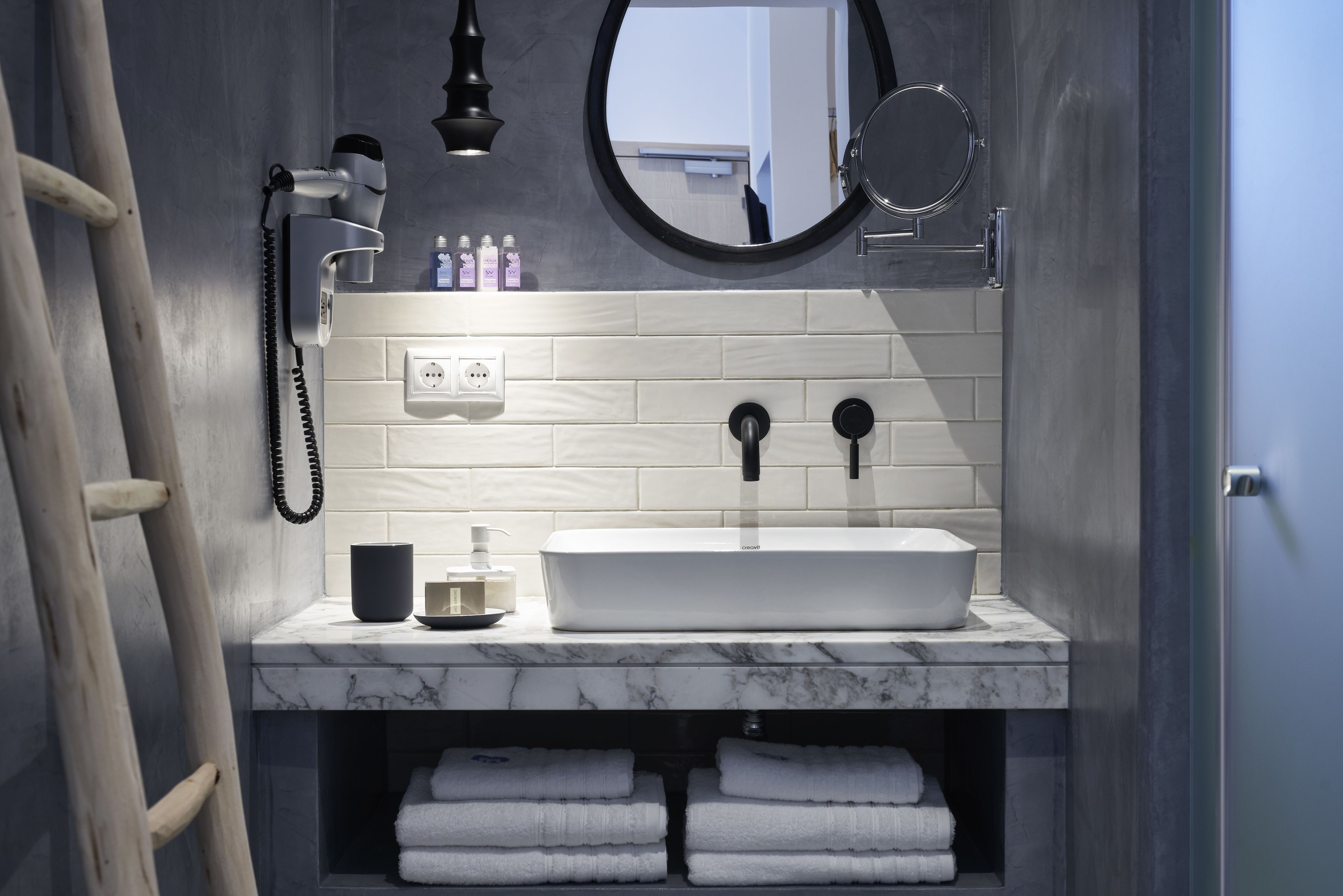Un baño con un lavabo blanco, un espejo redondo y un toallero con toallas dobladas.