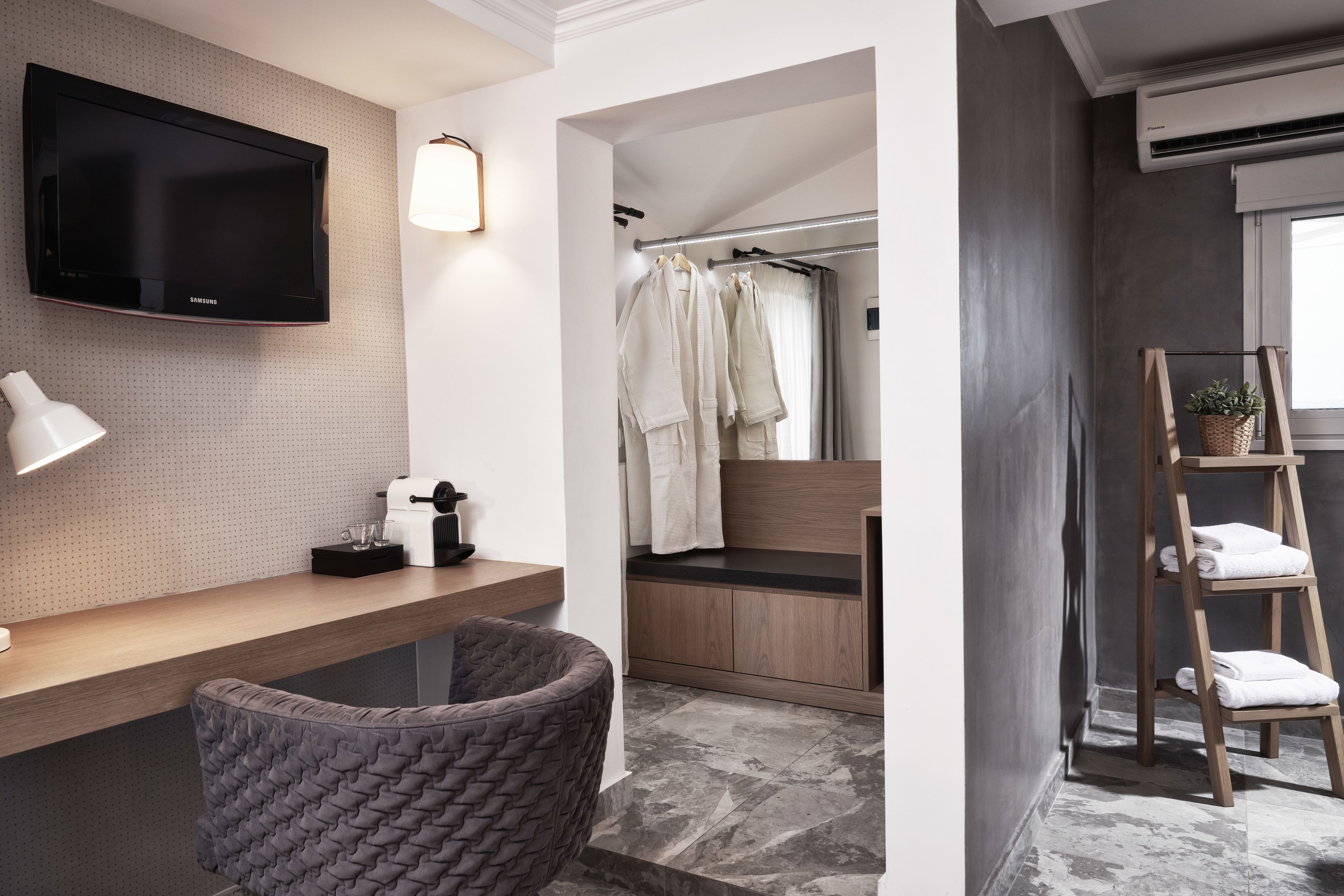 A modern bathroom with a mounted TV, a wooden vanity, a grey cushioned chair, a towel rack, and a wooden ladder with towels.