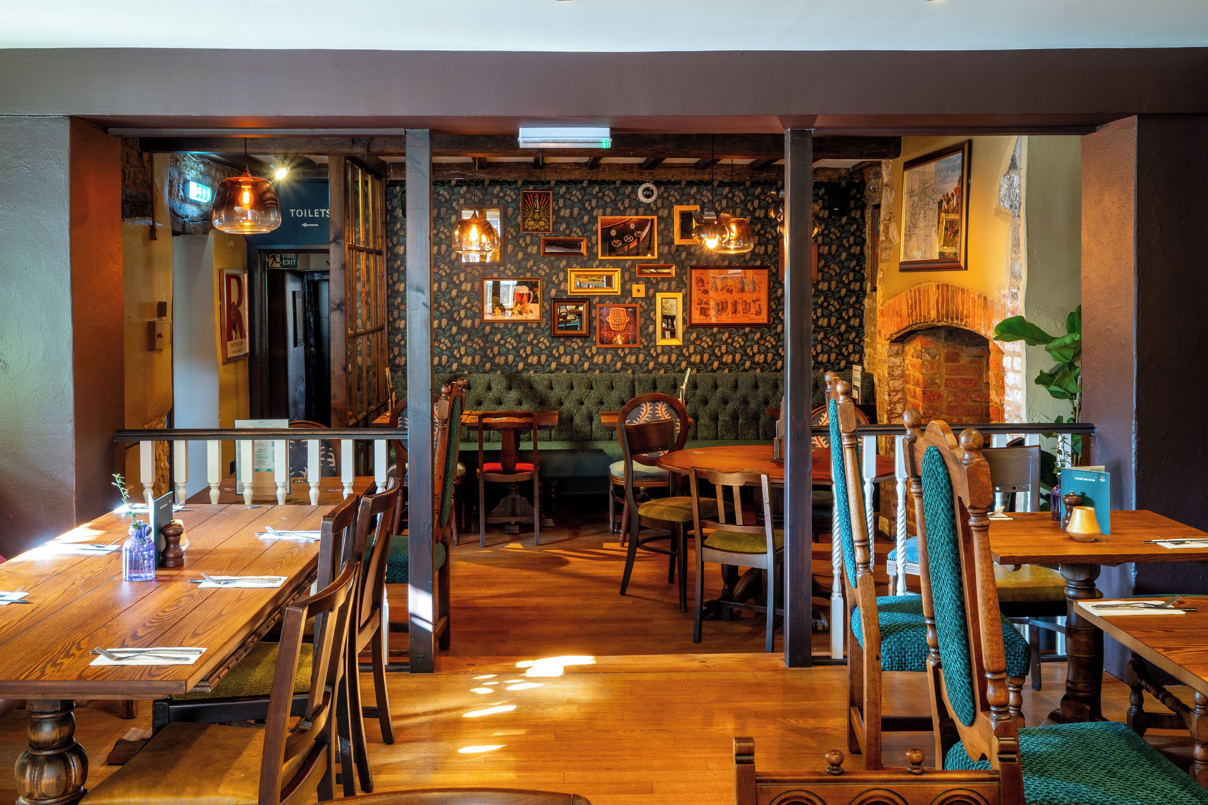 A restaurant interior with wooden tables and chairs, a patterned wall with framed pictures, and a brick fireplace.