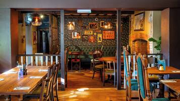 A restaurant interior with wooden tables and chairs, a patterned wall with framed pictures, and a brick fireplace.