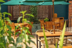 An outdoor seating area with wooden tables and chairs, green umbrellas, and a wicker wall.