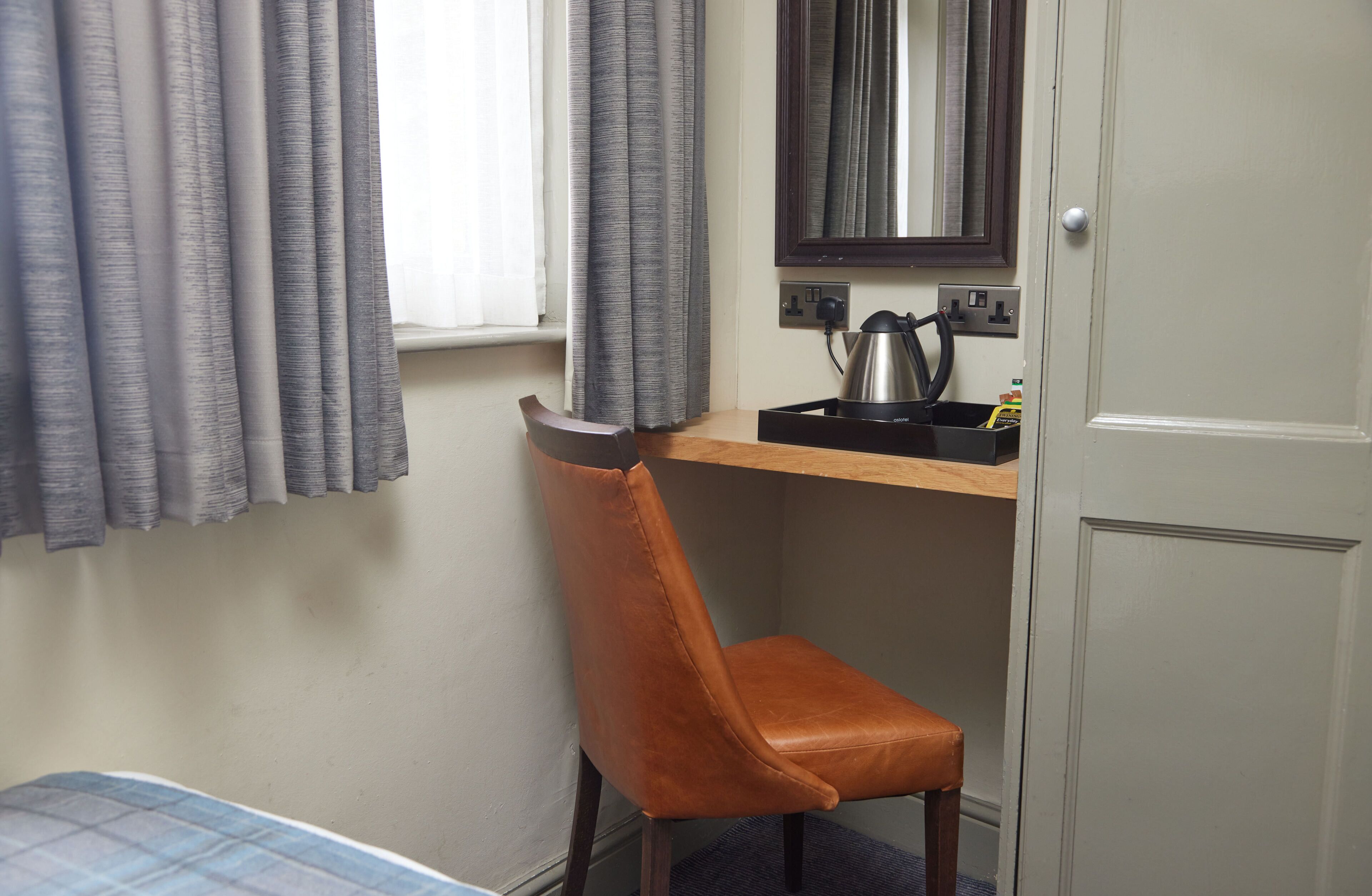 Una habitación pequeña con una cama, un sillón de cuero marrón, un escritorio de madera con un hervidor y un espejo.