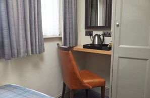 A small room with a bed, a brown leather chair, a wooden desk with a kettle, and a mirror.