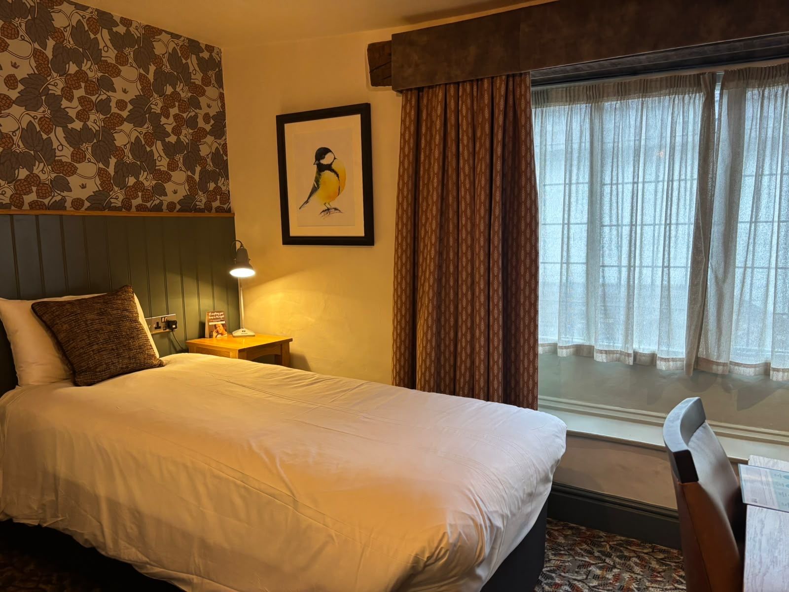 A hotel room with a large bed, a bedside table with a lamp, a framed picture on the wall, and a window with curtains.