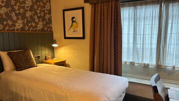 A hotel room with a large bed, a bedside table with a lamp, a framed picture on the wall, and a window with curtains.